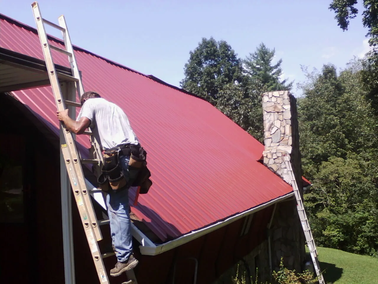 Roofer on a ladder beside a red metal roof and stone chimney — Soffit & Fascia Repair by Roofing Brothers in Limington