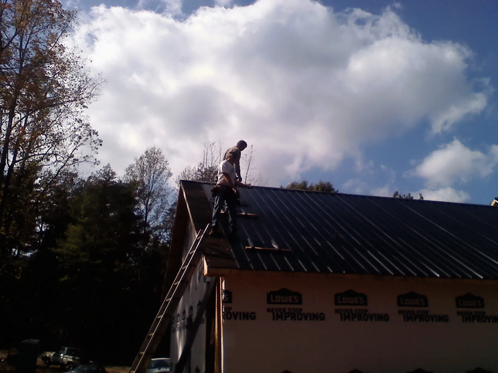Roofer fastening dark metal panels high against a cloudy sky, from a Roof Inspection job in Limington